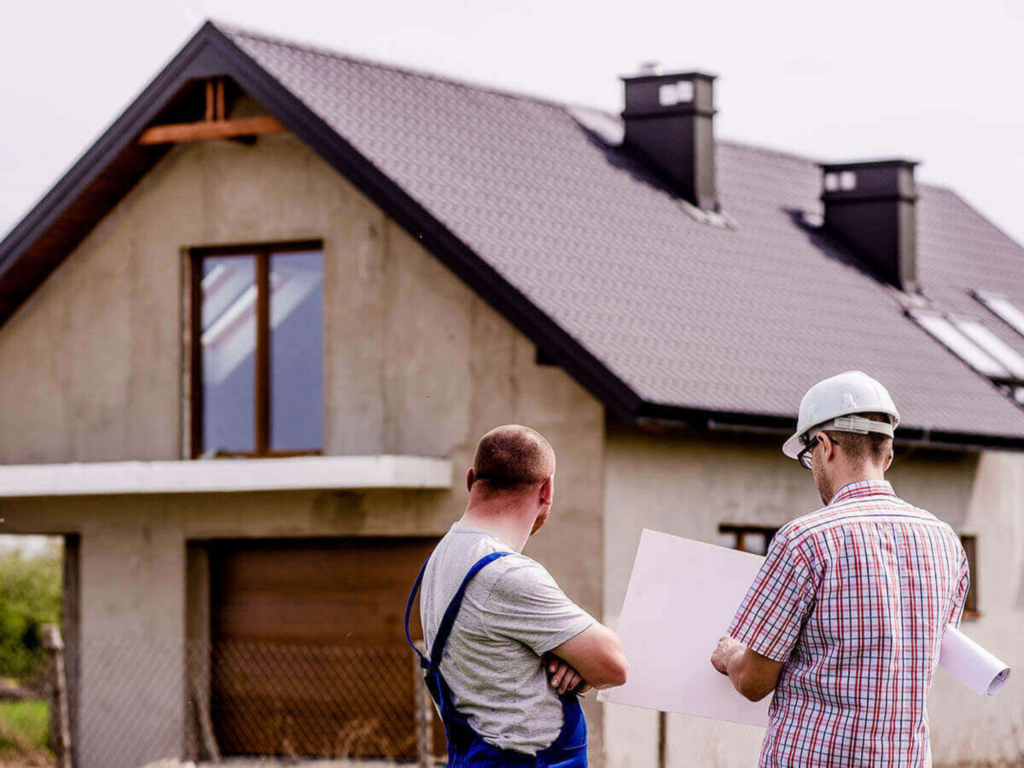 Two men in construction attire discussing a blueprint in front of an unfinished house with a gabled roof.