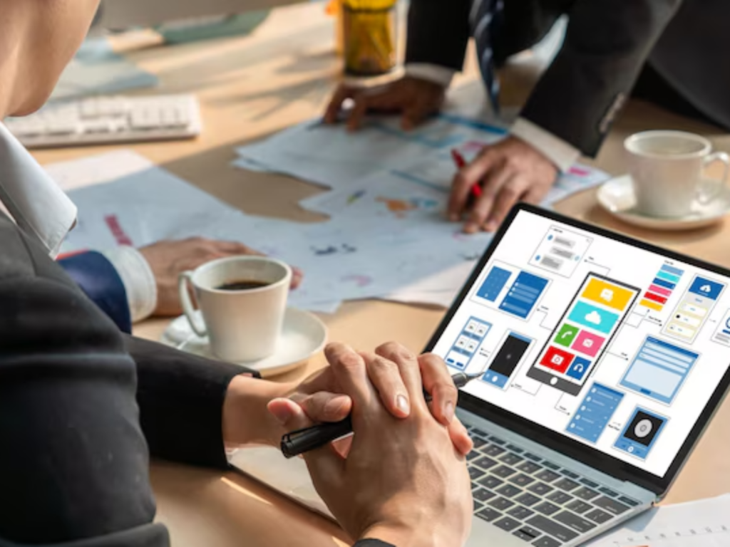 Person reviewing app design wireframes on a laptop at a desk with coffee cups and papers.