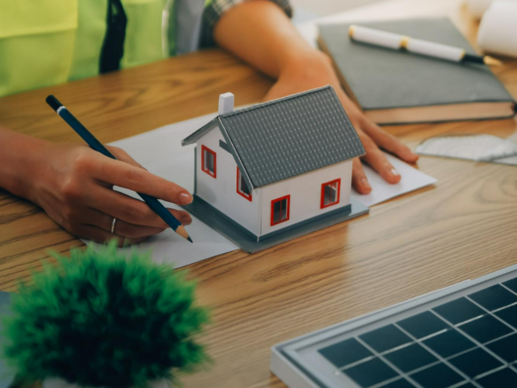 Person sketching near a small house model on a table, with a solar panel and various items in the background.